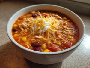 A close-up of a white bowl filled with 7 can chicken taco soup, topped with shredded cheese and sour cream.