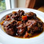 Close-up of tender brown stew chicken pieces coated in a rich, dark sauce with visible vegetable chunks.