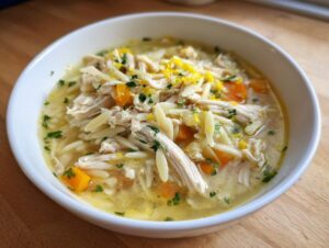 Close-up of a white bowl filled with amazing chicken orzo soup, featuring shredded chicken, carrots, and bright lemon zest.