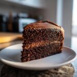 A close-up of a moist slice of amazing chocolate cake layered with rich frosting on a white plate.
