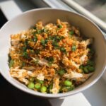 A close-up of a bowl containing crispy rice salad mixed with shredded chicken, bright green edamame, and topped with fresh herbs.