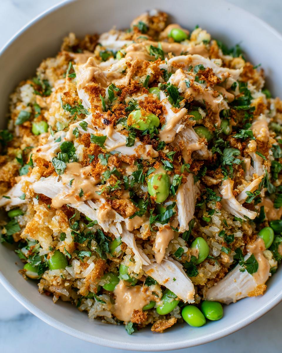 A bowl filled with crispy rice salad topped with shredded chicken, bright green edamame, and a creamy orange dressing.
