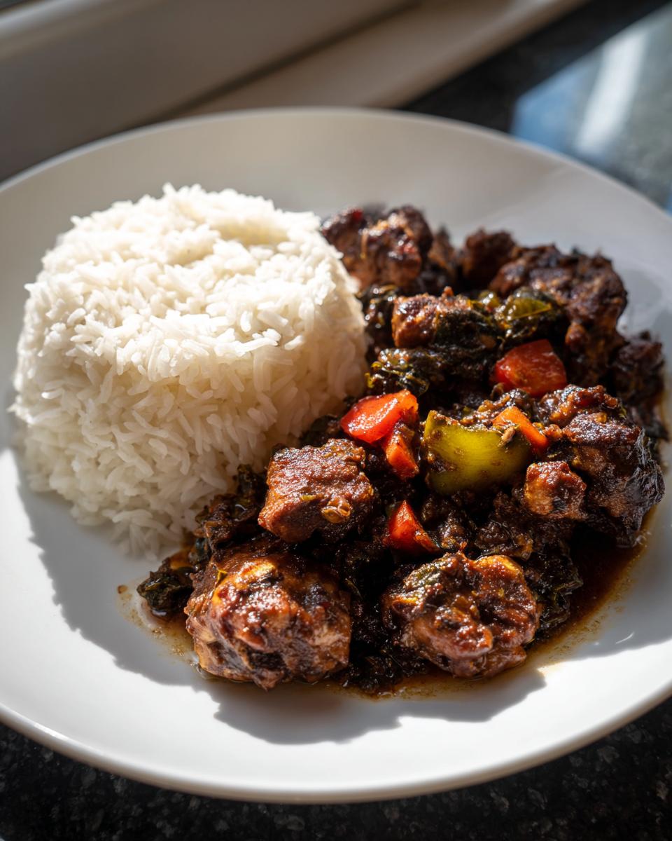 A close-up of rich brown stew chicken served next to a mound of fluffy white rice on a white plate.