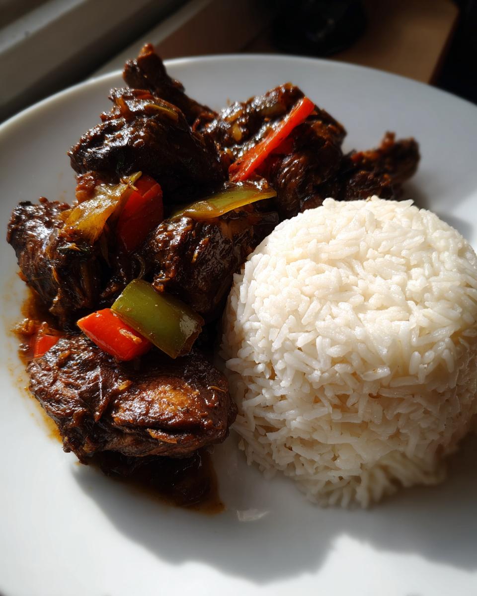 A plate featuring rich brown stew chicken with peppers next to a perfectly molded mound of white rice.