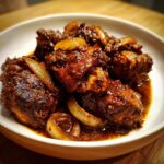 Close-up of rich, dark brown stew chicken jamaican crock pot pieces with caramelized onions in a white bowl.