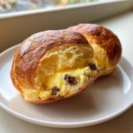 Two golden brown chocolate chip vanilla custard brioches, one cut open to show creamy yellow custard filling.