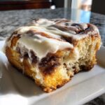 A close-up, tempting shot of a square slice of cinnamon roll cake topped with thick white icing.