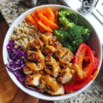 Close-up of a vibrant buddha bowl featuring chicken, quinoa, broccoli, carrots, and peanut sauce.