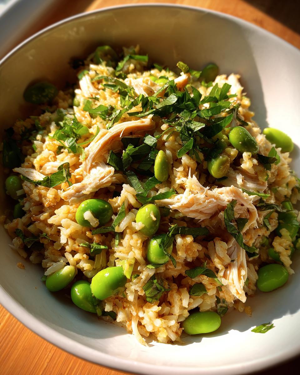 Close-up of a bowl containing crispy rice salad mixed with shredded chicken, bright green edamame beans, and fresh herbs.