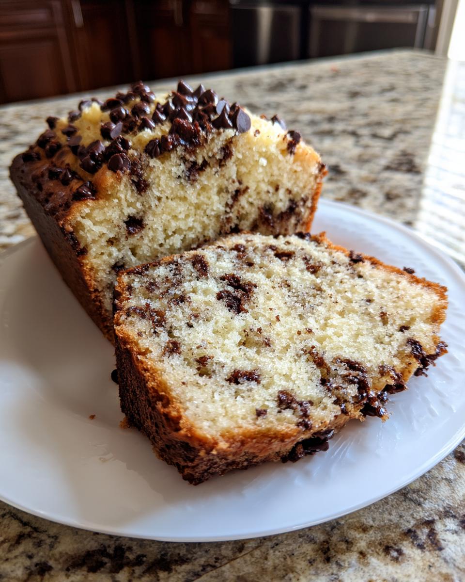 A sliced vanilla chocolate chip cake loaf, rich with chocolate chips and topped with more chips, sitting on a white plate.