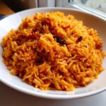 A close-up of fluffy, orange-red jollof rice nigerian served in a white bowl near a window.