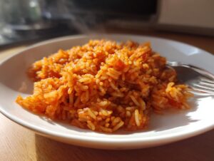 A close-up of a serving of vibrant, orange-red smoky jollof rice steaming slightly on a white plate next to a fork.