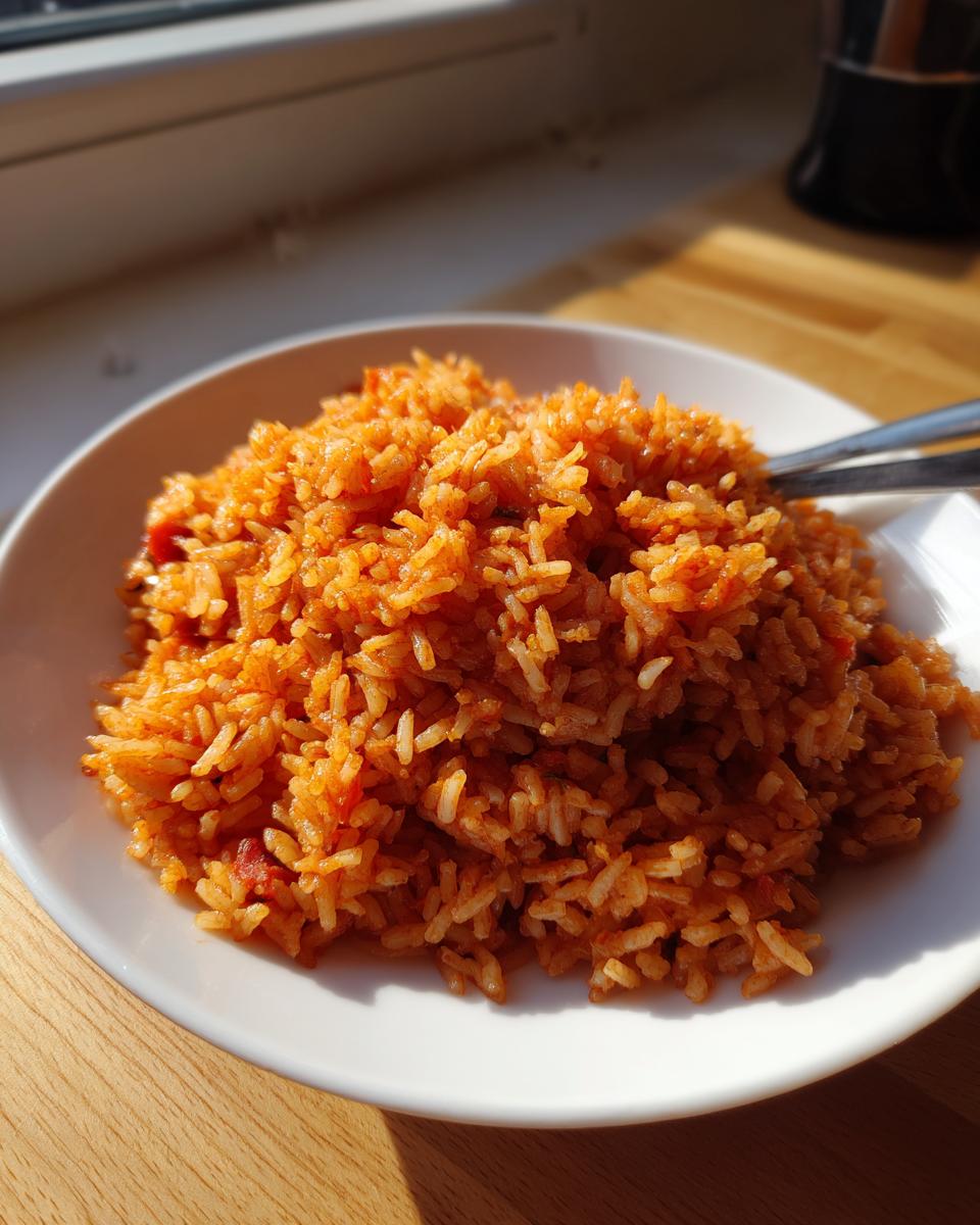 A generous serving of vibrant, orange-red jollof rice piled high on a white plate, illuminated by sunlight.