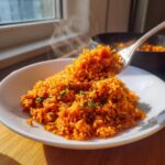A spoonful of steaming, vibrant orange jollof rice being lifted from a white bowl.
